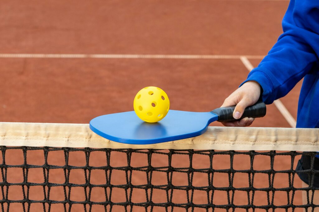 Player holds blue pickleball paddle with yellow ball by the net, symbolising that pickleball is popular with Malaysian punters.